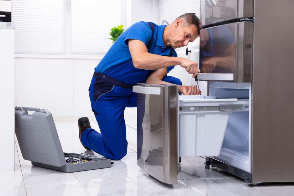 Mature Male Serviceman Repairing Refrigerator With Toolbox In Kitchen